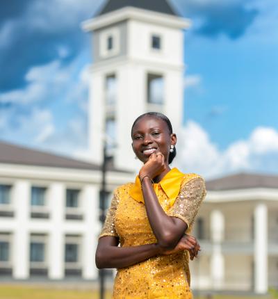 This is the image of a young lady standing and posing with her hand at her chin smiling.